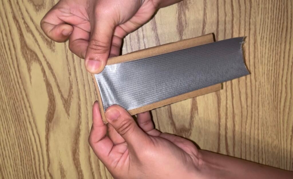 Hands wrapping a strip of silver duct tape around a piece of cardboard.