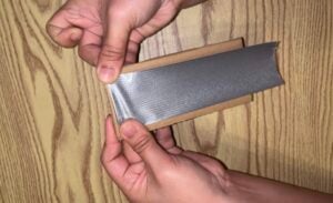 Hands wrapping a strip of silver duct tape around a piece of cardboard.