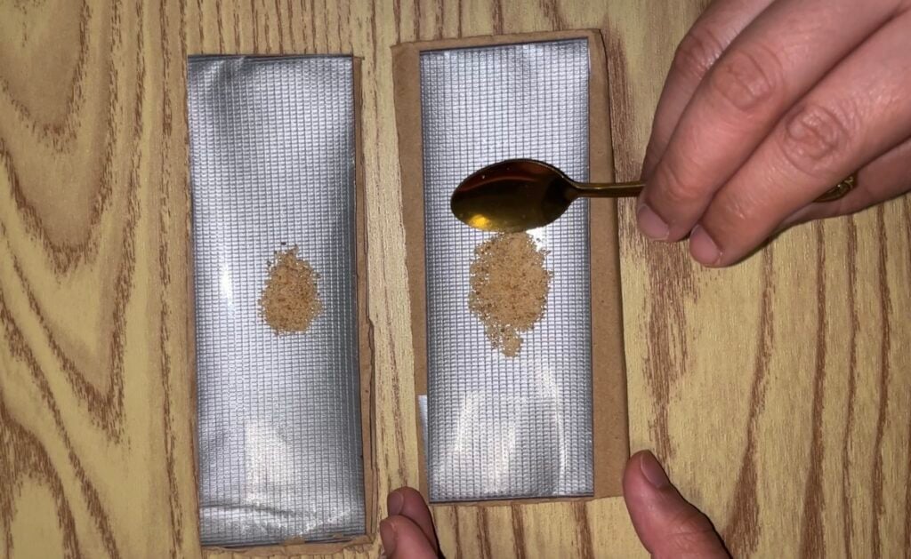 A hand uses a gold spoon to drop a small mound of brown powder onto a silver adhesive strip, next to an identical strip with powder already on it.