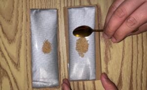 A hand uses a gold spoon to drop a small mound of brown powder onto a silver adhesive strip, next to an identical strip with powder already on it.