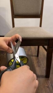 Hands cutting a piece of silver duct tape with scissors in front of a wooden dining chair.