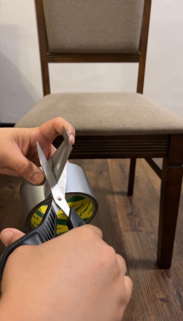 Hands cutting a piece of silver duct tape with scissors in front of a wooden dining chair.