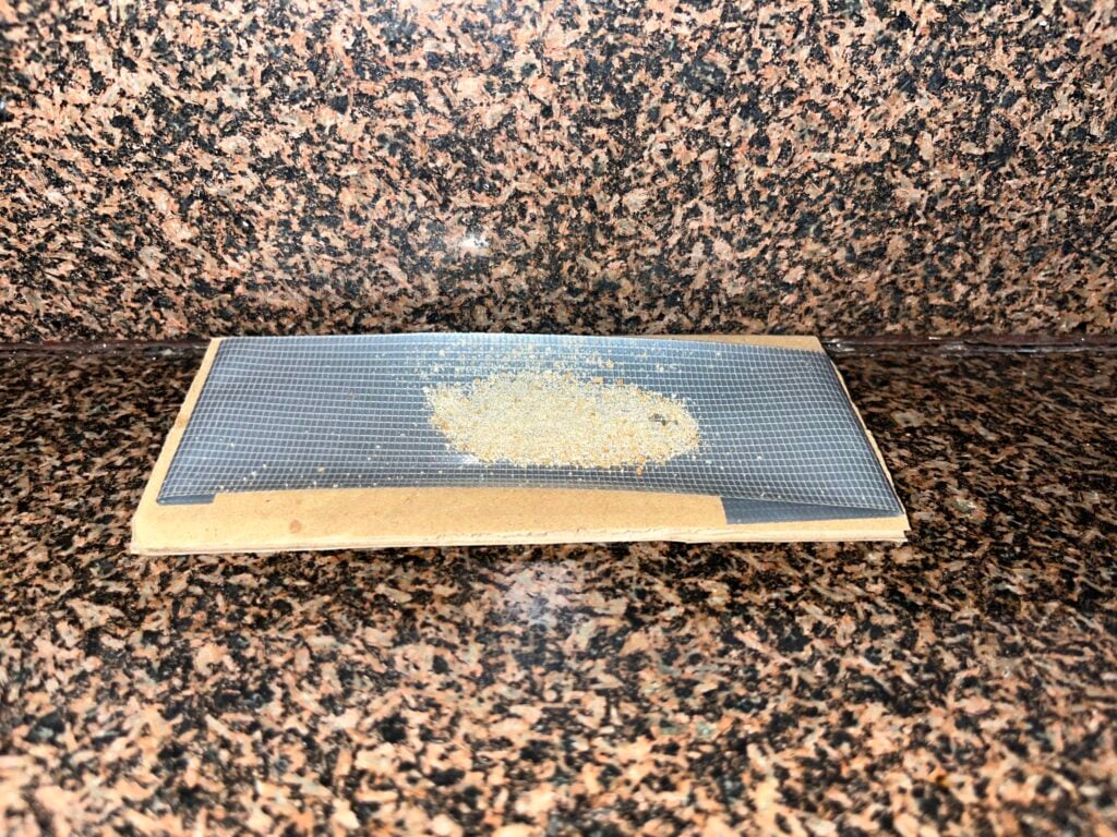 A small pile of light-colored, granular substance sits on a piece of dark, fine-mesh screen resting on a cardboard base on a granite countertop.