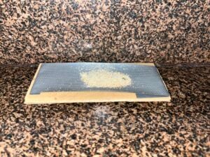 A small pile of light-colored, granular substance sits on a piece of dark, fine-mesh screen resting on a cardboard base on a granite countertop.