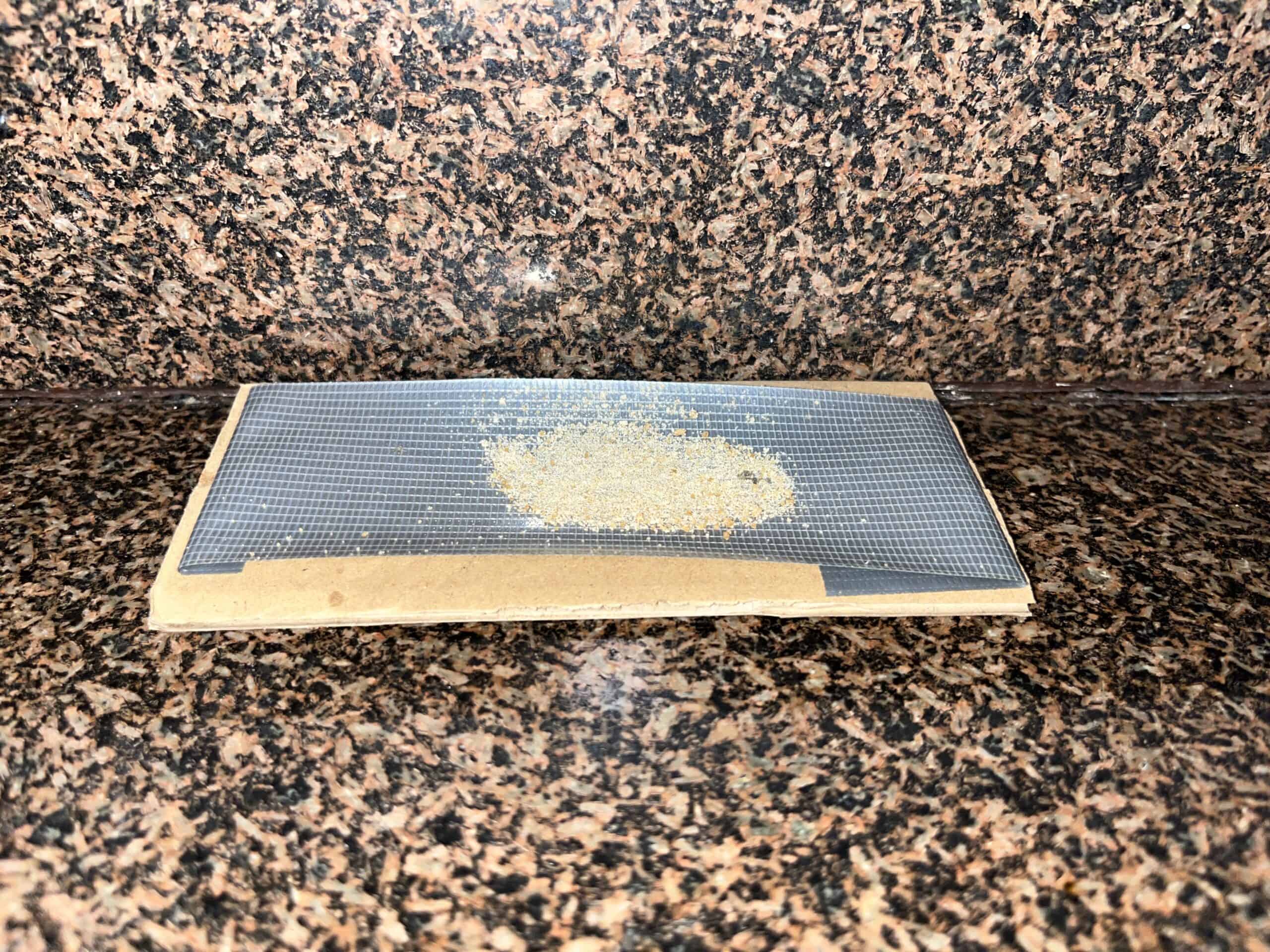 A small pile of light-colored, granular substance sits on a piece of dark, fine-mesh screen resting on a cardboard base on a granite countertop.